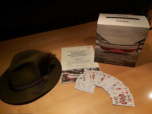 Auf einem Tisch liegen anlässlich einer Doppelkopf-Spendenaktion ein aufgefächertes Kartenspiel, eine Seenotretter-Sammelbox mit Begleitschreiben sowie ein Jägerhut.